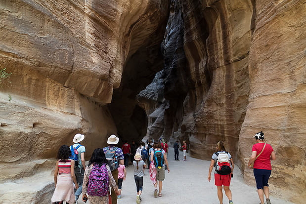 A group of people walk through a narrow, towering canyon with layered rock walls, wearing backpacks and casual attire, suggesting a hiking or sightseeing expedition.